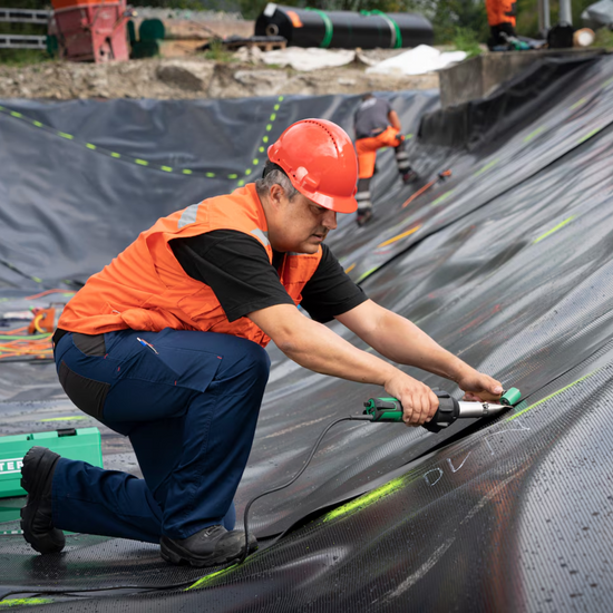 Worker using green handheld geomembrane welder to seam black HDPE pond liner on site