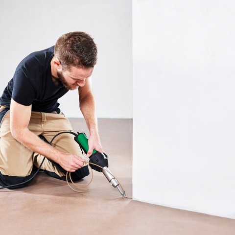 Professional worker using green corded hot air heat gun on flooring edge near wall baseboard.