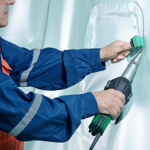 Worker using green hot air welder to seam white PVC tarp with roller rod