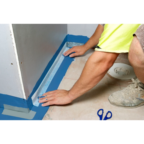 Worker applying blue painter's masking tape along wall-floor edge for clean, precise paint lines.