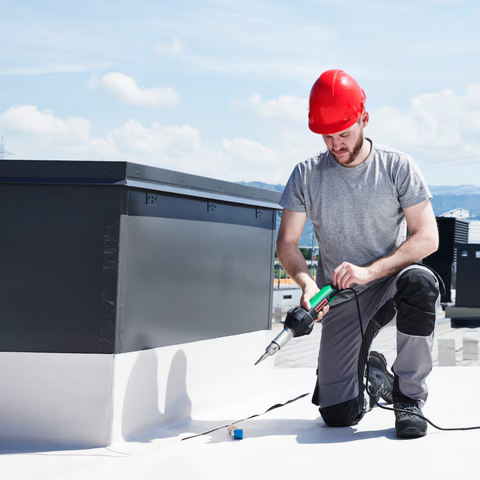 Worker using green handheld hot air roofing welder on flat membrane seams, professional rooftop tool.