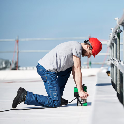 Professional roofer in red hard hat using green handheld roof edge trimmer on flat rooftop